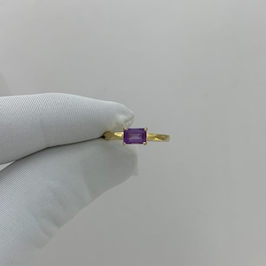 May include: A gold ring featuring a rectangular, deep purple gemstone. The ring's band is a simple, polished gold, and the gemstone is set in a classic, minimalist style. The ring is shown against a plain white background.