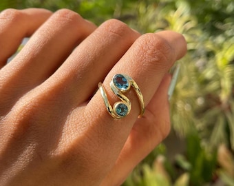 Puede incluir: Un anillo de oro tipo bypass con dos piedras preciosas azules redondas engastadas en bisel. La banda tiene un diseño abierto y superpuesto que se curva alrededor del dedo, mostrando el metal dorado pulido y el color azul vivo de las piedras.