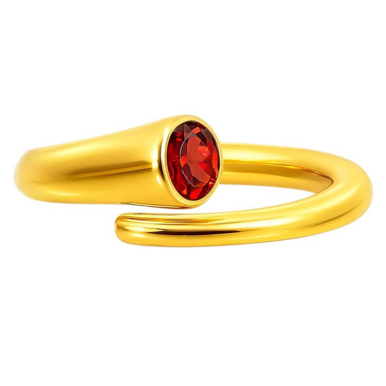 May include: A gold-colored ring with a vibrant red oval-shaped gemstone. The ring has a smooth, polished finish and a modern, minimalist design. The gemstone is set in a bezel setting, adding to the ring's elegant appearance. A piece of jewelry.
