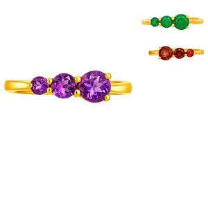 May include: A gold ring with three purple gemstones. The ring has a simple band and the gemstones are graduated in size. Two other rings are also shown, one with green gemstones and one with red gemstones.