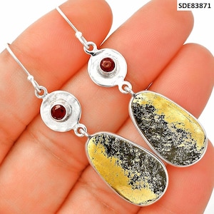 May include: Silver dangle earrings with a red gemstone set in a hammered silver circle. The earrings feature teardrop-shaped pendants with a black and gold speckled design. The earrings are held in a hand.