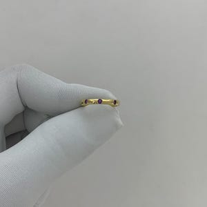 May include: A gold ring with three small, round, purple gemstones. The ring is held against a white background. The ring's band is a smooth, polished gold.
