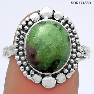 May include: A silver ring featuring a large, oval-shaped green gemstone with red inclusions. The gemstone is set in a decorative bezel with a textured border of small silver beads and larger silver accents. The ring is shown on a finger.