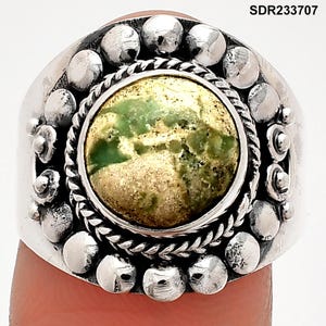 May include: Silver ring with a large, round, green and gold stone center. The ring has a textured silver band with decorative silver beads around the stone. The ring is on a finger.