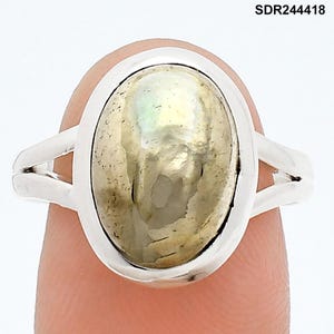 Puede incluir: Un anillo de plata con una piedra preciosa ovalada e iridiscente. La piedra preciosa tiene un color claro y nacarado con sutiles tonos verdes y amarillos. La banda del anillo se divide en dos líneas paralelas a cada lado de la piedra.