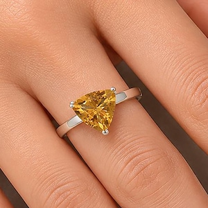 Op de afbeelding: Een zilverkleurige metalen ring met een enkele, gefacetteerde, driehoekig geslepen gele edelsteen in een zetting met drie pootjes. De ringband is glad en gepolijst, wat het strakke ontwerp benadrukt en de levendige goudgele steen accentueert.