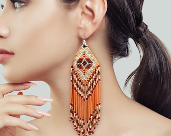 May include: A long, dangling earring crafted from small beads in shades of orange, white, brown, and red. The earring features a diamond-shaped top section with a geometric pattern and a long fringe made of orange and multi-colored beaded strands.