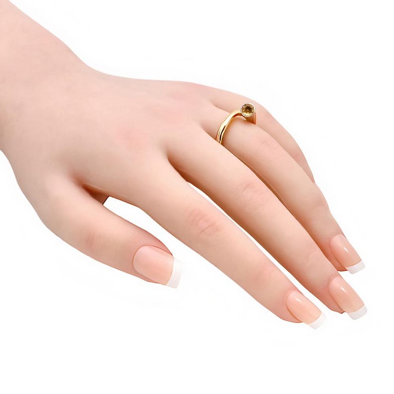 May include: A gold ring with a small, round, amber-colored gemstone. The ring is on a hand with manicured nails. The ring's band curves elegantly, and the gemstone is set in a simple, classic style. The ring is a piece of jewelry.