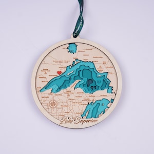 May include: A wooden ornament shaped like Lake Superior, with a layered, 3D design showing the depth of the lake. The ornament is painted in shades of blue and green, with a red heart marking the approximate location of the Apostle Islands. The ornament is labeled "Lake Superior" at the bottom.