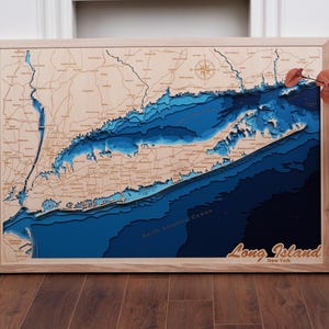 Peut inclure: Une carte en bois de Long Island, New York, avec un relief 3D détaillé de la terre et de l'océan environnant. La carte est encadrée de bois clair et porte l'inscription "Long Island New York" dans une police stylisée.