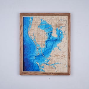 May include: A framed, layered wood map of Tampa Bay, Florida, USA. The map features detailed blue water depths and a light brown wooden frame. The text "Tampa Bay" is visible.