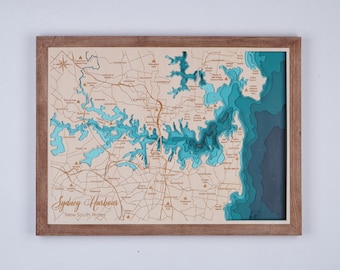 May include: A wooden map of Sydney Harbour, New South Wales, with a brown frame. The map is layered with blue and teal shades to represent the water. The map is titled "Sydney Harbour, New South Wales".