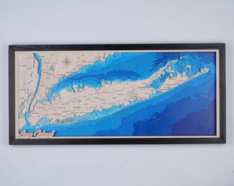 May include: A framed wooden map of Long Island, New York, with a black frame. The map features a detailed depiction of the island's topography, with varying shades of blue representing the depth of the surrounding waters. The landmass is a light tan color.