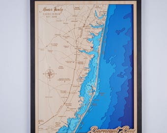 May include: A wooden map of Barnegat Bay, New Jersey, with a black frame. The map is layered and shows the depth of the water in shades of blue. The text "Mouser Family Shore House Est. 2001" is in the top left corner. The text "Barnegat Bay Ocean County, NJ" is in the bottom right corner.