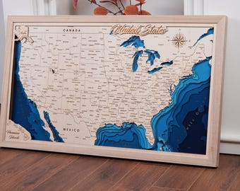 May include: A wooden map of the United States with blue 3D layers representing the ocean. The map is framed in a light wood frame and includes the text "United States" and "Canada" on the top of the map.