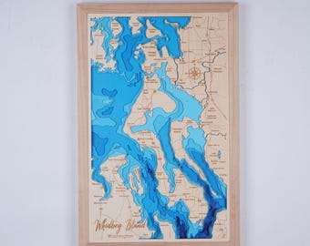 May include: A wooden map of Whidbey Island, Washington, with blue water depths carved into the wood. The map is framed in a light wood frame.