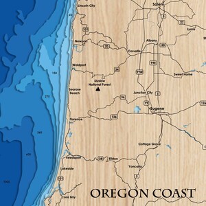 Puede incluir: Un mapa de la costa de Oreg&oacute;n con las principales ciudades y autopistas etiquetadas. El mapa est&aacute; impreso sobre un fondo de madera.