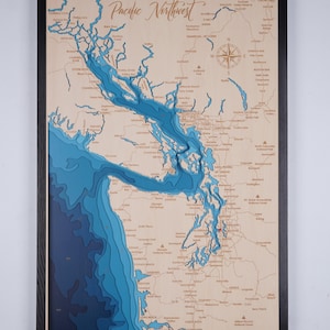 Pode incluir: Um mapa de madeira emoldurado do noroeste do Pacífico, apresentando um relevo 3D detalhado da costa e das vias navegáveis em tons de azul. O mapa inclui nomes de lugares e uma rosa dos ventos, com o título "Pacific Northwest" em escrita elegante.