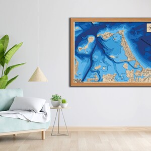 May include: A framed wooden map of Hull, Massachusetts, with a blue and white color scheme depicting the depth of the water. The map is framed in a light wood frame and includes a compass rose in the top right corner.