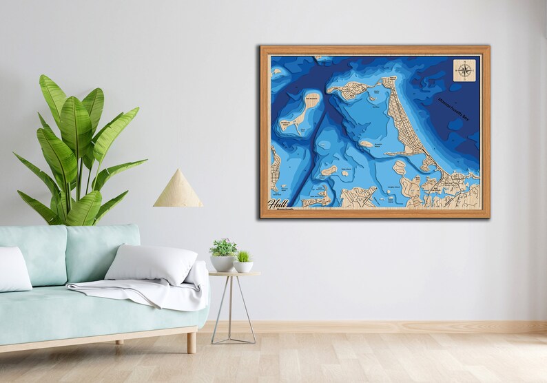 May include: A framed wooden map of Hull, Massachusetts, with a blue and white color scheme depicting the depth of the water. The map is framed in a light wood frame and includes a compass rose in the top right corner.