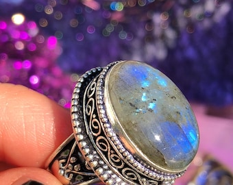 May include: A silver ring with an oval-shaped labradorite gemstone. The ring has intricate filigree details and a beaded border around the stone.