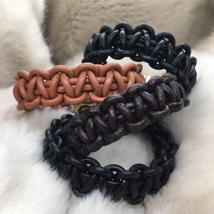 May include: Three braided leather bracelets in black, dark brown, and tan. Each bracelet features a thick, interlocking woven pattern created from rounded leather cords, showcasing a consistent, repetitive knot design throughout the entire band.