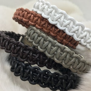 Peut inclure: Une collection de quatre bracelets tressés en blanc, brun clair, gris et noir. Chaque bracelet présente un design en cordon épais et tissé avec une surface texturée, empilés ensemble pour montrer les variations de couleur distinctes.