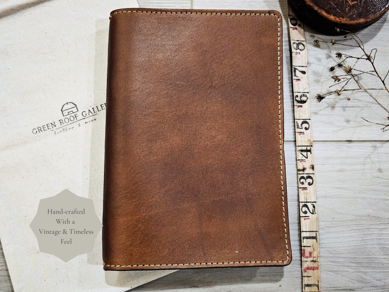 Puede incluir: Un cuaderno de cuero marr&oacute;n con un borde cosido y el texto "GREEN ROOF GALLERY" en la portada. El cuaderno est&aacute; hecho a mano con un toque vintage y atemporal.