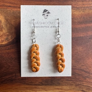 May include: A pair of handmade earrings featuring a braided bread design. The earrings are made of polymer clay and are on silver hooks. The card they are displayed on reads "The Mushroom Circle Handcrafted Jewelry".