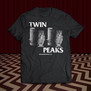 May include: Black t-shirt featuring a white graphic of three owls perched on tree stumps. The text "Twin Peaks" is below the owls, with the words "Washington U.S.A." underneath.