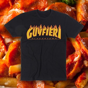 May include: Black t-shirt with the text "GUY FIERI FLAVORTOWN" in yellow and orange flame-style lettering. The shirt is displayed against a background of pizza and french fries.