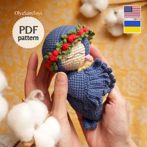 Op de afbeelding: Een handgemaakte blauwe gehaakte pop met een bloemenkrans. De pop heeft een lachend gezicht en houdt een PDF-patroon vast. De pop is ongeveer 10 cm hoog. De afbeelding bevat ook katoenbollen.