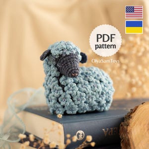 Può includere: Una pecora all'uncinetto fatta a mano, nei toni del blu e del grigio. La pecora ha un corpo strutturato e soffice, e un viso e orecchie grigio scuro. Il testo "PDF pattern" e il nome "OlyaSamToys" sono inclusi.