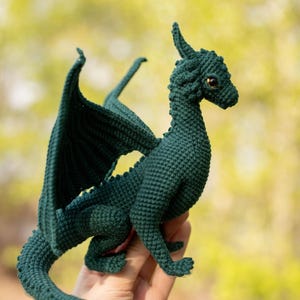 May include: A dark green crochet dragon, featuring detailed wings, horns, and a long tail. The dragon has golden eyes and is posed dynamically. The background is a soft, blurred green. The dragon is made with a crochet technique.