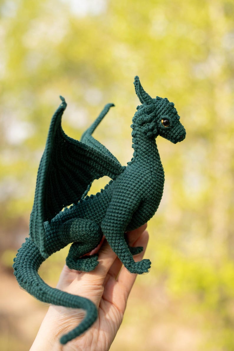 May include: A dark green crochet dragon, featuring detailed wings, horns, and a long tail. The dragon has golden eyes and is posed dynamically. The background is a soft, blurred green. The dragon is made with a crochet technique.