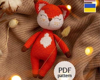 May include: A handmade orange and white crocheted fox doll with brown accents. The fox has a white diamond shape on its chest and is lying on a brown knitted surface. The image includes the text "PDF pattern" and "OlyaSamToys".