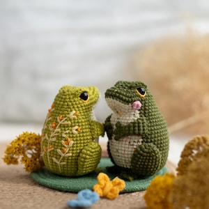 Puede incluir: Dos figuras de rana de ganchillo hechas a mano. Una rana es de color verde claro con bordados florales naranjas, la otra es de color verde oscuro con una mejilla rosa. Se enfrentan, sobre una estera verde, con peque&ntilde;os elementos decorativos.