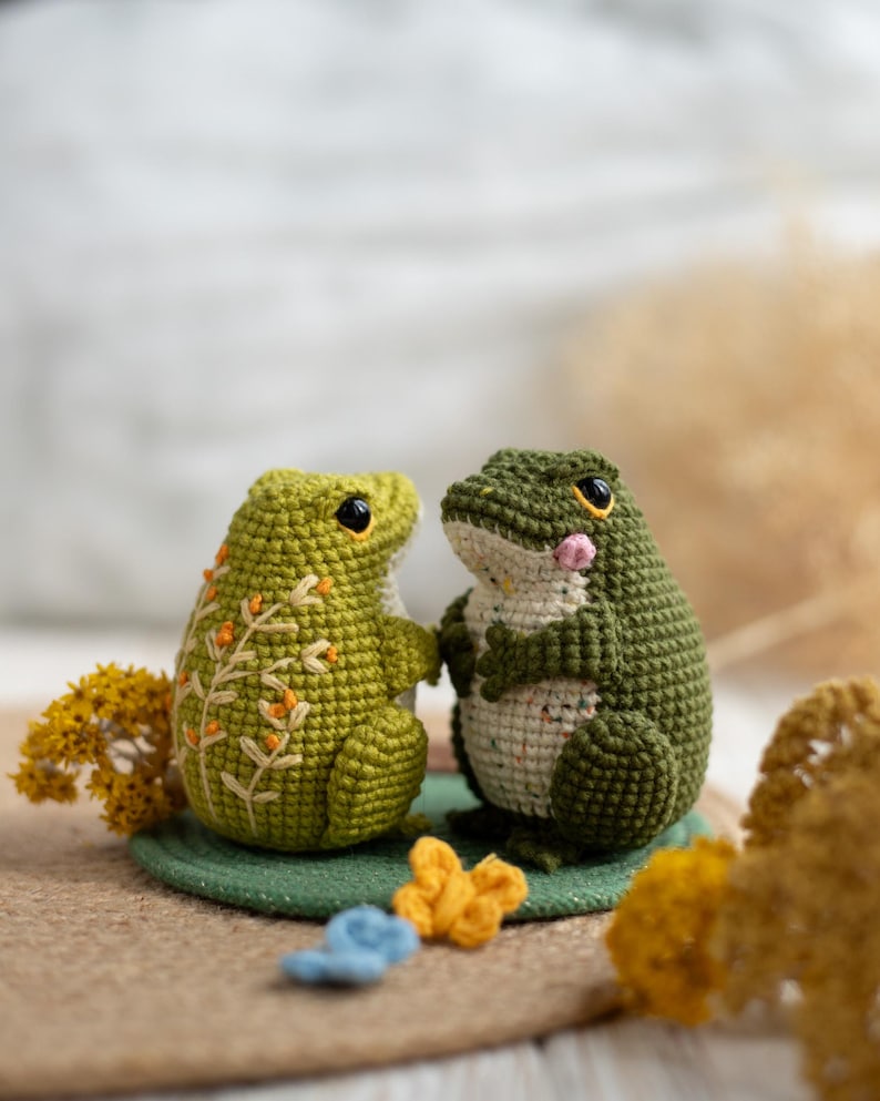 Puede incluir: Dos figuras de rana de ganchillo hechas a mano. Una rana es de color verde claro con bordados florales naranjas, la otra es de color verde oscuro con una mejilla rosa. Se enfrentan, sobre una estera verde, con peque&ntilde;os elementos decorativos.