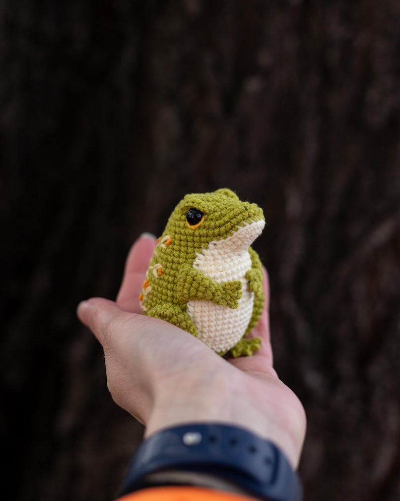 Puede incluir: Una rana de ganchillo hecha a mano en tonos verdes y crema, con detalles en naranja. La rana tiene ojos negros y se sostiene en una mano abierta. La rana es un art&iacute;culo artesanal caprichoso.