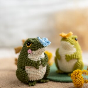 Puede incluir: Dos figuras de rana de ganchillo hechas a mano. Una rana es verde con una flor azul en la cabeza, la otra es verde con una flor amarilla. Las ranas tienen vientres blancos y est&aacute;n sobre una superficie marr&oacute;n.