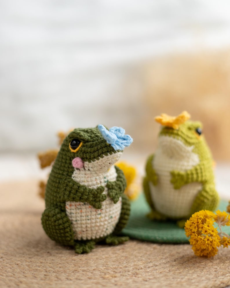 Puede incluir: Dos figuras de rana de ganchillo hechas a mano. Una rana es verde con una flor azul en la cabeza, la otra es verde con una flor amarilla. Las ranas tienen vientres blancos y est&aacute;n sobre una superficie marr&oacute;n.