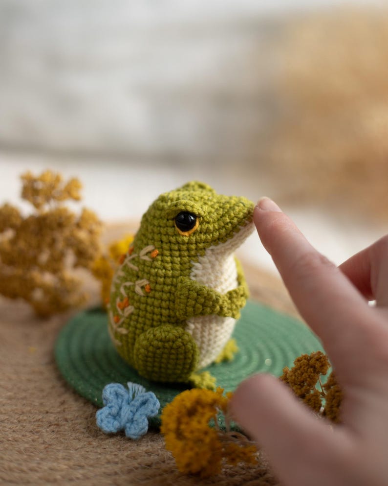 Puede incluir: Una rana de ganchillo hecha a mano en tonos verdes y crema, con ojos negros y detalles en naranja. La rana est&aacute; sentada sobre una estera circular verde, con un dedo tocando su nariz. Flores secas decorativas y una flor azul de ganchillo en el fondo.