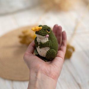 Puede incluir: Una rana de ganchillo hecha a mano en tonos verdes y crema, con una flor amarilla en la cabeza. La rana tiene ojos negros y mejillas rosadas. Este amigurumi caprichoso es un art&iacute;culo decorativo encantador.