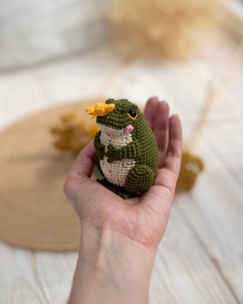 Puede incluir: Una rana de ganchillo hecha a mano en tonos verdes y crema, con una flor amarilla en la cabeza. La rana tiene ojos negros y mejillas rosadas. Este amigurumi caprichoso es un art&iacute;culo decorativo encantador.