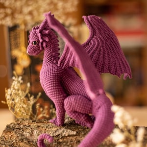 May include: A handmade, purple crochet dragon figure with detailed wings, a long tail, and small, round eyes. The dragon is posed on a piece of wood, showcasing its textured, scale-like body. The background is blurred, highlighting the dragon.