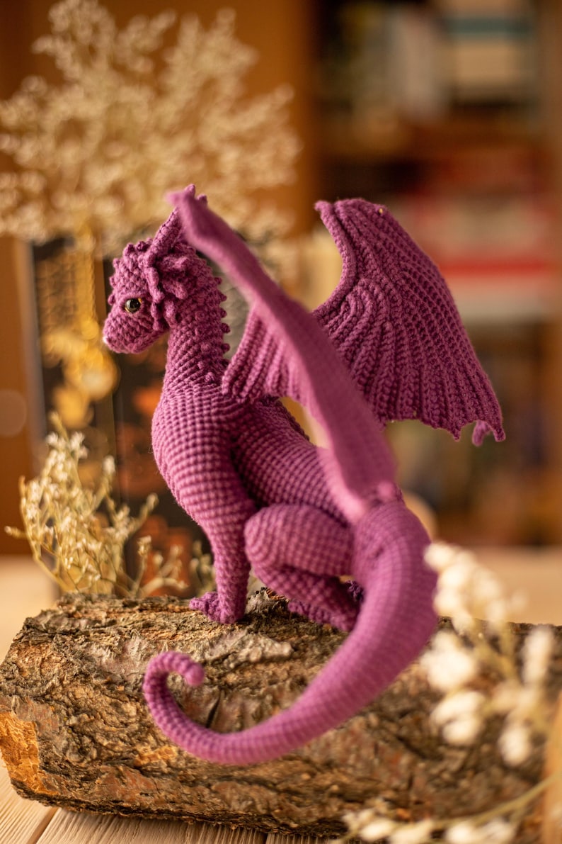 May include: A handmade, purple crochet dragon figure with detailed wings, a long tail, and small, round eyes. The dragon is posed on a piece of wood, showcasing its textured, scale-like body. The background is blurred, highlighting the dragon.