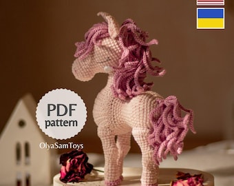 May include: A crocheted unicorn with a pink body and a flowing, curly pink mane and tail. The unicorn has a single horn and is standing on a decorative base. The image includes the text "PDF pattern" and the name "OlyaSamToys".