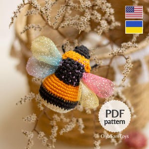 May include: A handmade crochet bee with orange, black, and yellow stripes, and iridescent wings. The bee is embellished with small purple beads. The image includes the text "PDF pattern" and "OlyaSamToys".