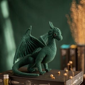 May include: A handmade, dark green crochet dragon with detailed wings and a textured body. The dragon is posed on a stack of decorative books with gold accents. A small bottle of glitter sits nearby.
