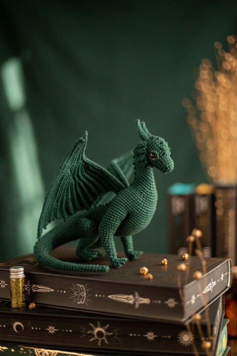 May include: A handmade, dark green crochet dragon with detailed wings and a textured body. The dragon is posed on a stack of decorative books with gold accents. A small bottle of glitter sits nearby.